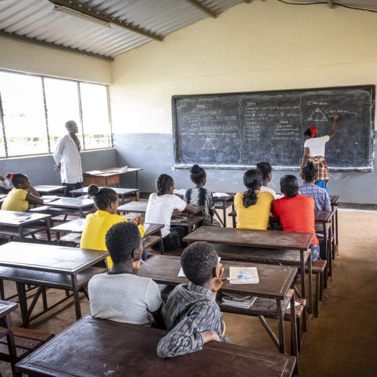 Schüler und Lehrer beobachten, wie ein Mädchen in einem hellen, belebten Klassenzimmer auf eine Kreidetafel schreibt.