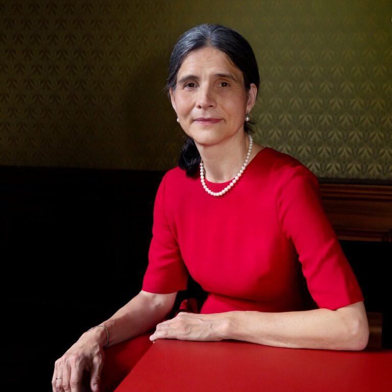 Grauhaarige Frau in rotem Kleid, Perlenkette, sitzt an einem roten Tisch mit Blick nach vorn.