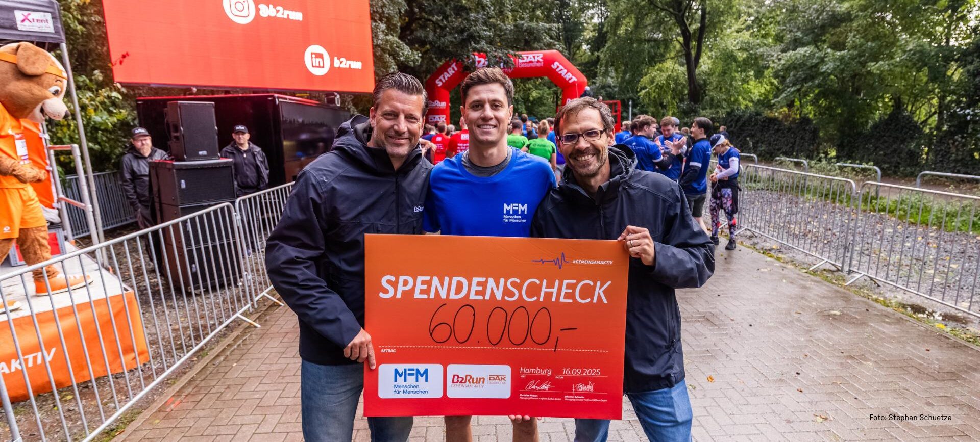 Drei lächelnde Männer, die bei einer NGO-Veranstaltung im Freien einen Spendenscheck in Höhe von 60 000 Euro in der Hand halten.