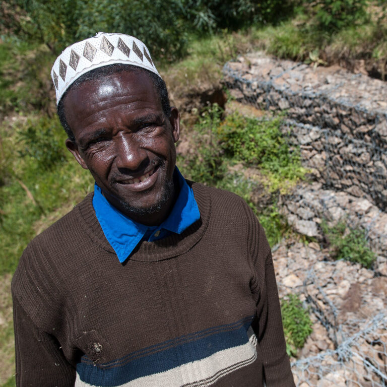 Ein lächelnder Mann in einem braunen Pullover und einer gemusterten Mütze steht im Freien zwischen Felsen und Grünpflanzen.