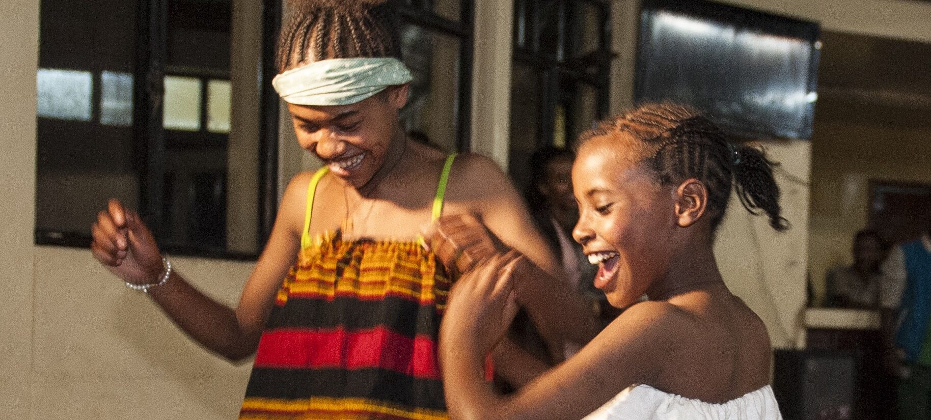 Zwei lächelnde Mädchen mit geflochtenen Haaren tanzen in farbenfrohen Kleidern im Haus, fröhlich und voller Leben.