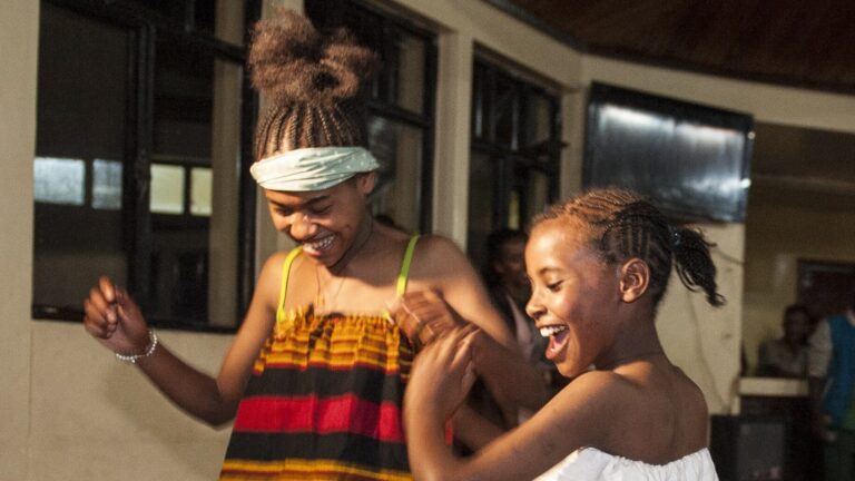 Lächelnde Mädchen in farbenfroher, traditioneller Kleidung tanzen zusammen in einem Haus.