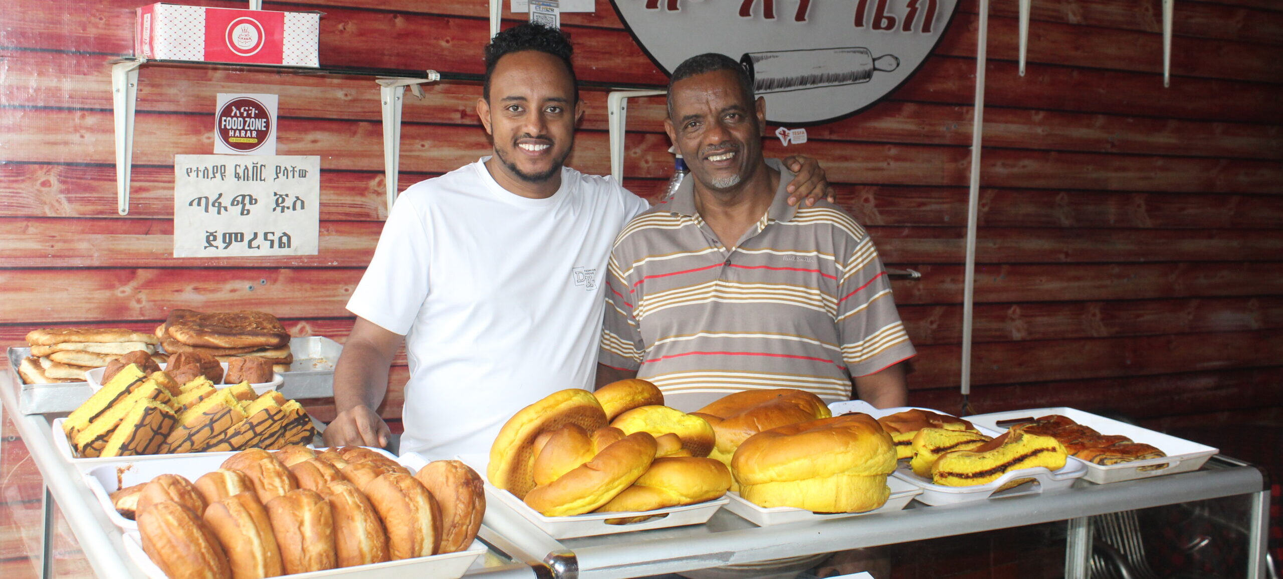Zwei Männer stehen lächelnd hinter einer Bäckertheke mit verschiedenen Brotsorten und Gebäck, die für die Spendenunterstützung stehen.