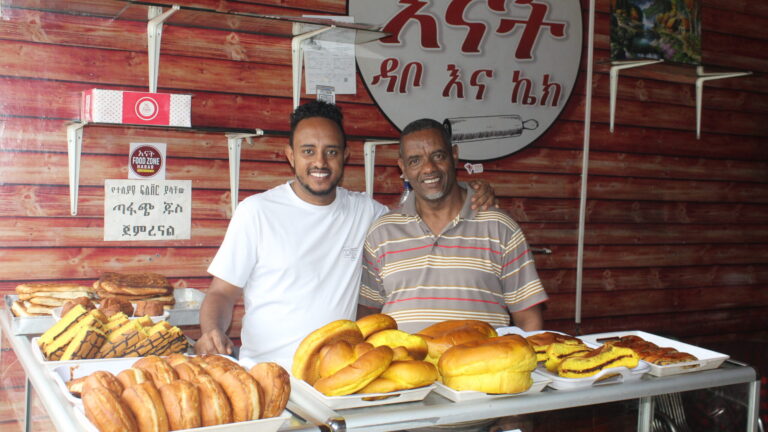Zwei lächelnde Männer stehen hinter einem Tisch mit verschiedenen Brotsorten in einer Bäckerei mit holzgetäfelten Wänden.