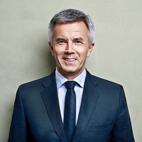 Ein lächelnder Mann in dunklem Anzug und Krawatte steht vor einer hellen Wand, die den Auftrag unserer NRO darstellt.