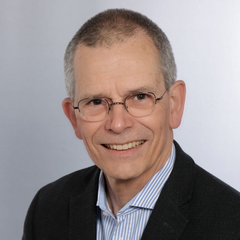 Freundlicher älterer Mann mit Brille und Anzugjacke, lächelnd vor einem schlichten hellen Hintergrund.