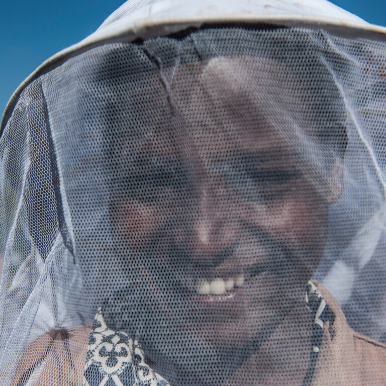 Eine Person in einem Imkerschleier lächelt, während sie draußen unter einem klaren blauen Himmel steht.