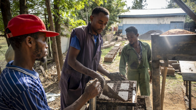 Drei Männer, die unter Bäumen mit einer Handpresse Ziegelsteine herstellen.
