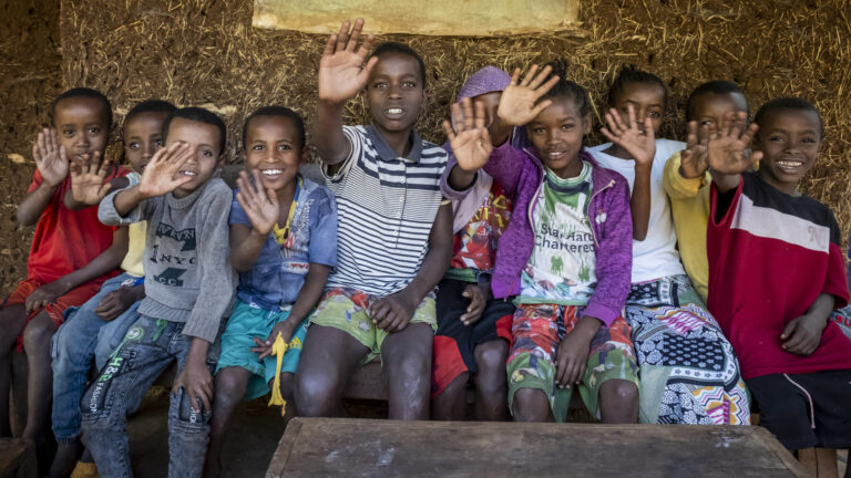Eine Gruppe von Kindern sitzt auf einer Bank. Sie heben freudig ihre Hand in die Höhe.