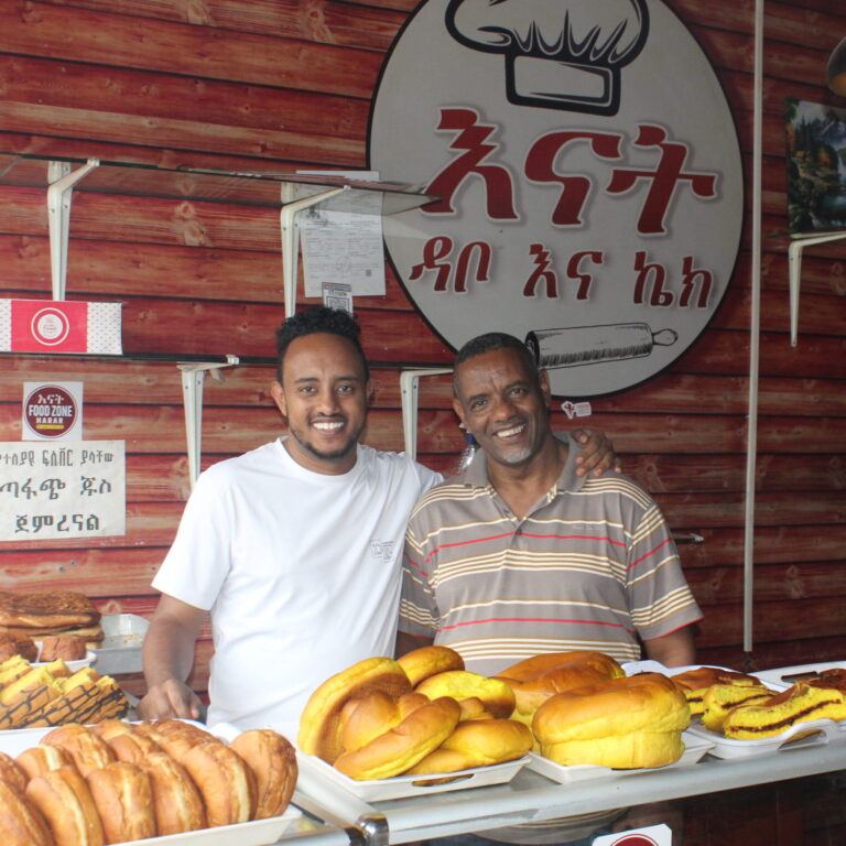 Zwei Männer stehen lächelnd hinter Tabletts mit Brot und äthiopischem Gewürzpesto in einer Bäckerei, um Spenden anzubieten.