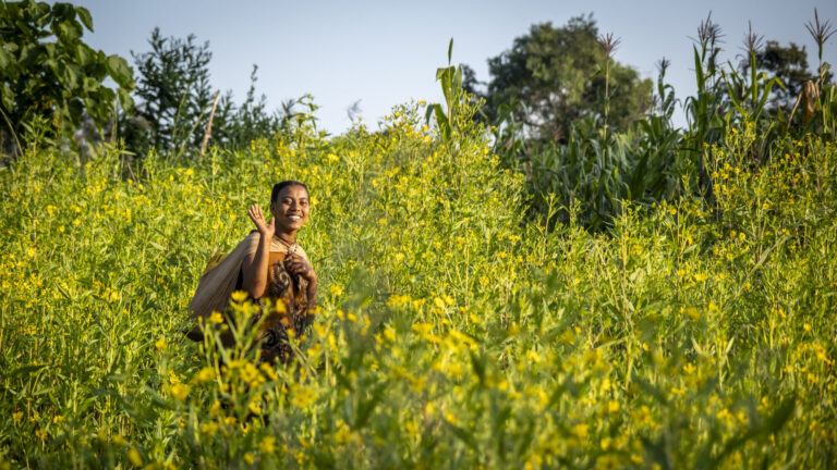 Eine Frau marschiert über ein Feld mit gelben Blumen. Sie trägt einen Wasserkanister auf dem Rücken.