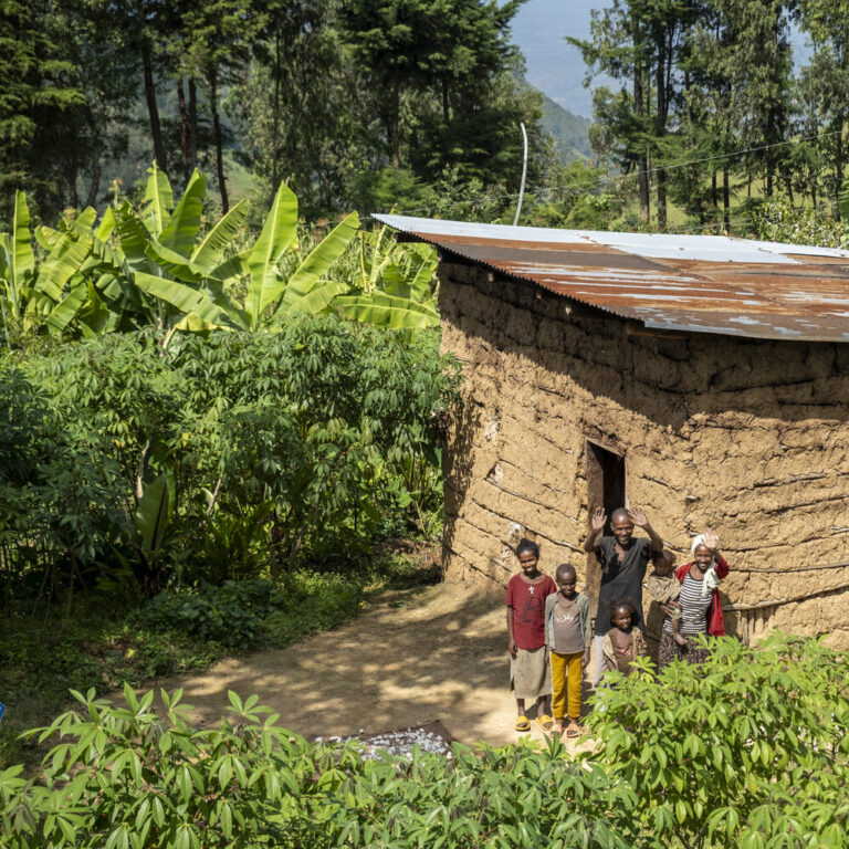 Eine Familie steht vor ihrem Wohnhaus, das umgeben von Bäumen und anderen Pflanzen ist.