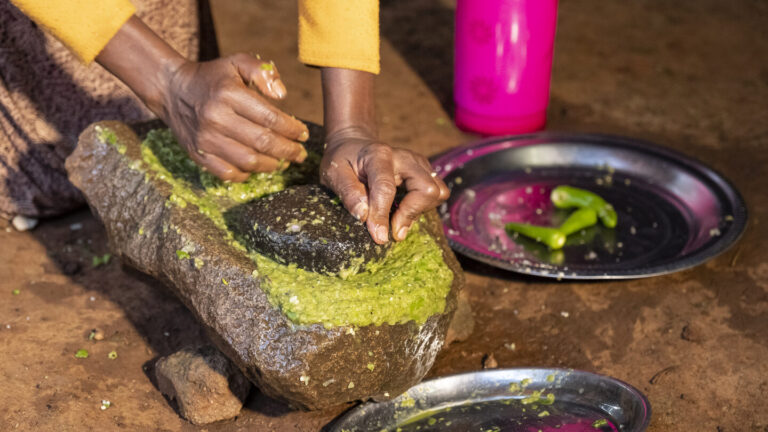 Hände, die grüne Chilis auf einem Stein zerkleinern.