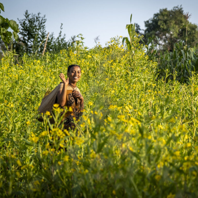 Eine Frau marschiert über ein Feld mit gelben Blumen. Sie trägt einen Wasserkanister auf dem Rücken.