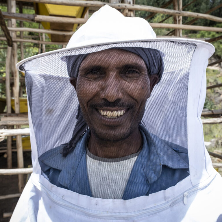Ein Imker in seinem Imkeranzug. Im Hintergrund sind Bienenkörbe.