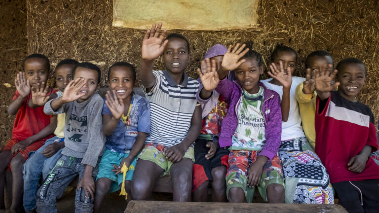 Eine Gruppe von Kindern sitzt auf einer Bank. Sie heben freudig ihre Hand in die Höhe.