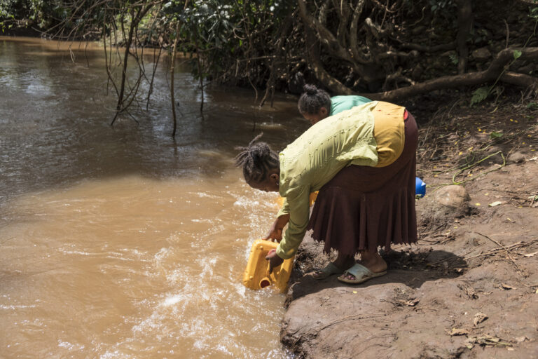 Eine Frau bückt sich und holt Wasser aus einem braunen fließenden Fluss.