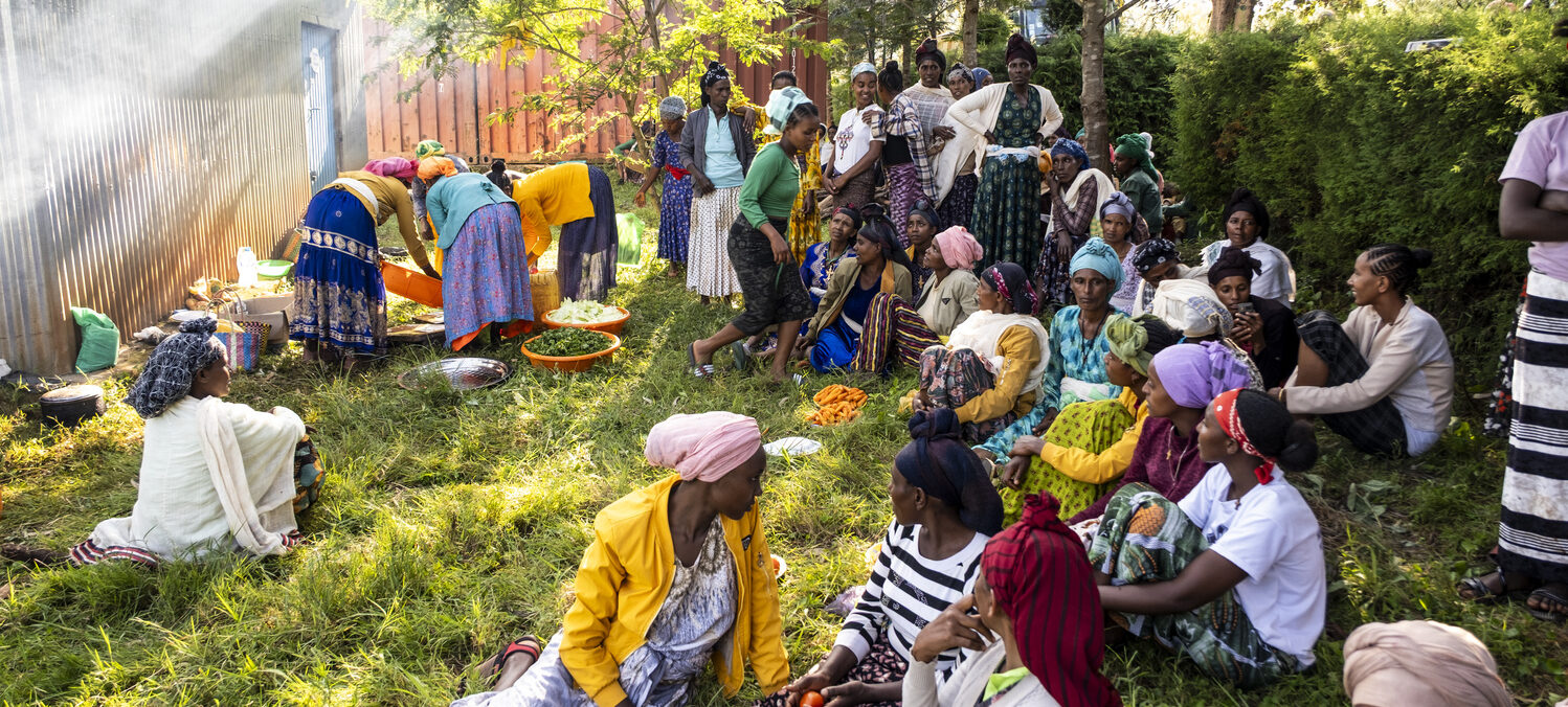 Eine Gruppe von Frauen mit bunten Kleidern sitzen im Grünen und unterhalten sich.