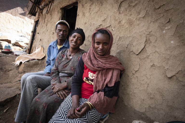 Eine Familie die vor ihrem Haus sitzt. und in die Kamera schaut. Yerom sitzt in der Mitte, ihre Mann links und ihre Tochter rechts von ihr.