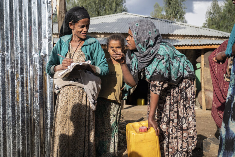 Zwei Frauen die miteinander reden. Eine frau hat einen Wasserkanister an ihrem Bein stehen, die andere hält einen Stift und Zettel in der hand.