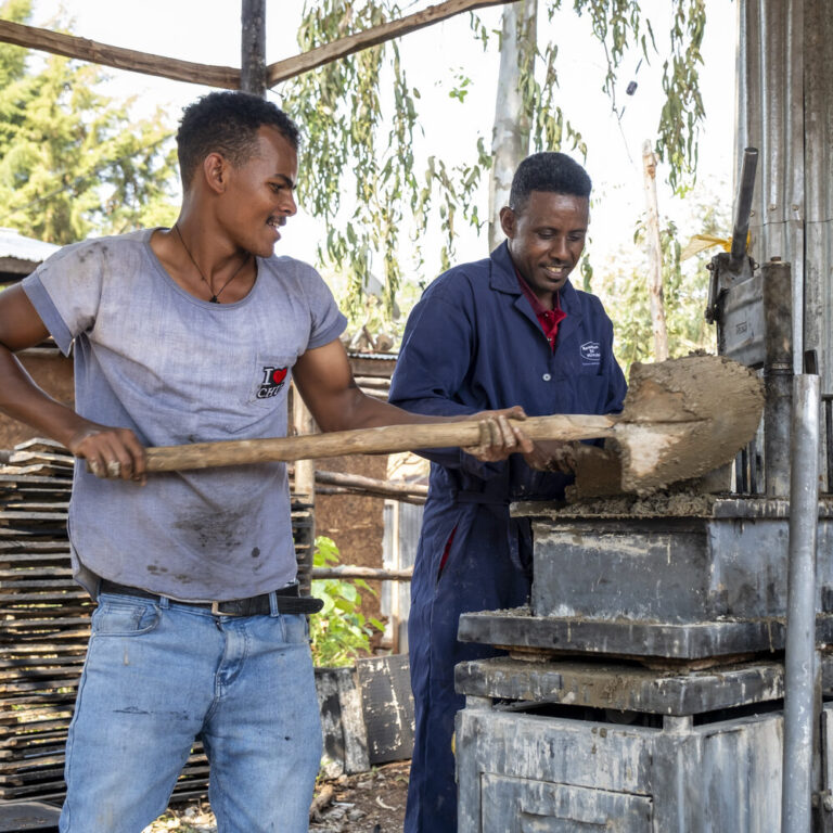 Drei Männer schaufeln Lehm und stellen Ziegel her, während sie im Sonnenlicht lächelnd zusammenarbeiten.