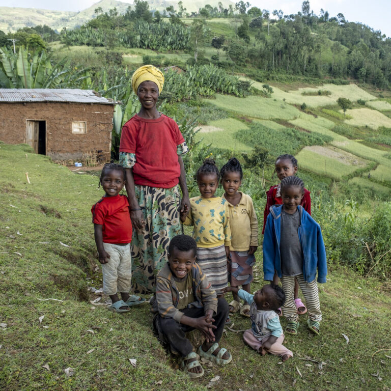 Eine Frau und sieben Kinder stehen dicht beieinander auf einer Wiese, im Hintergrund ist Ackerland zu sehen.