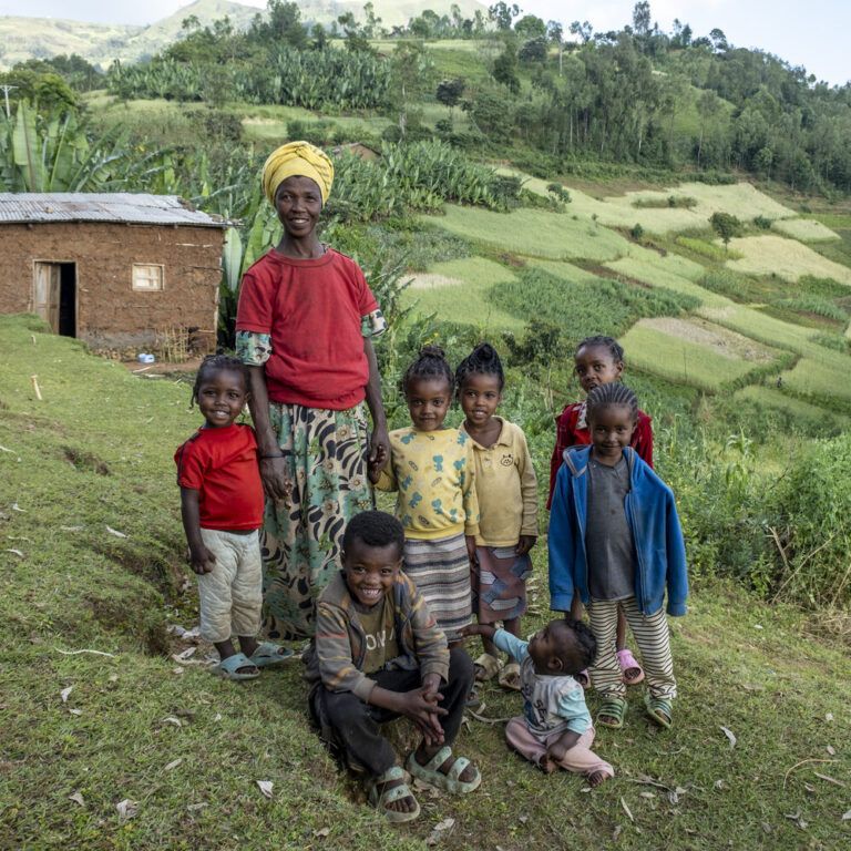 Eine Frau und sieben Kinder stehen dicht beieinander auf einer Wiese, im Hintergrund ist Ackerland zu sehen.