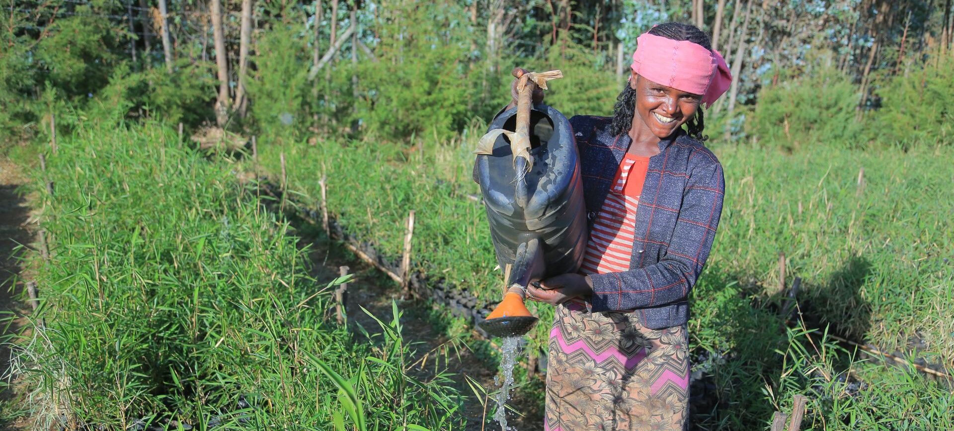 Eine Frau die lächelt und mit einer Gießkanne Pflanzen gießt.