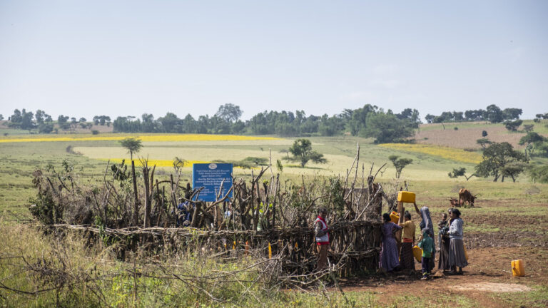 Frauen stehen Schlange um zur sauberen Wasserstelle zu kommen. Man sieht die mit Stöcken beschützte Stelle von außen.