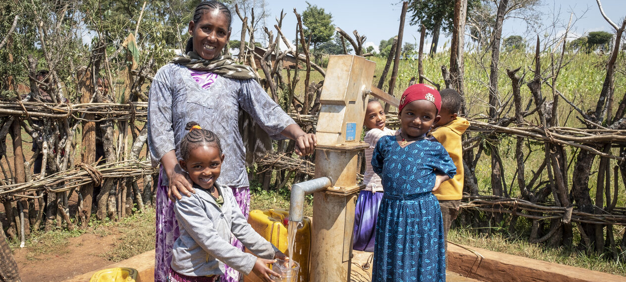 Eine Mutter steht mit ihren Kindern an einem Wasserbrunnen und lächelt in die Kamera.