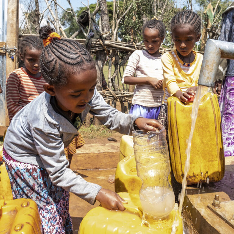 Vier Mädchen stehen am Wasserbrunne und füllen Wasser in Kanister.