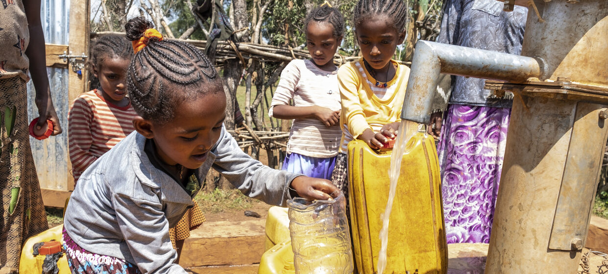 Vier Mädchen stehen am Wasserbrunne und füllen Wasser in Kanister.