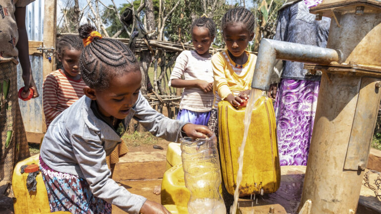Vier Mädchen stehen am Wasserbrunne und füllen Wasser in Kanister.