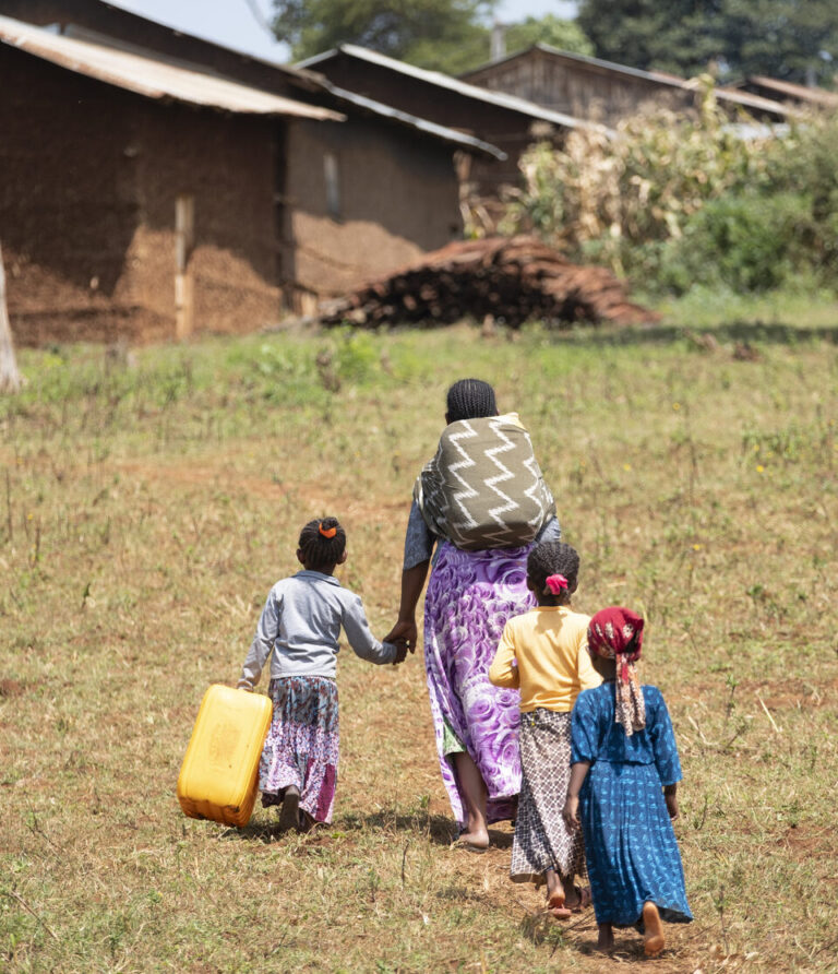 Frau geht mit ihren drei Kindern Weg entlang mit Wasserkanister auf dem Rücken.