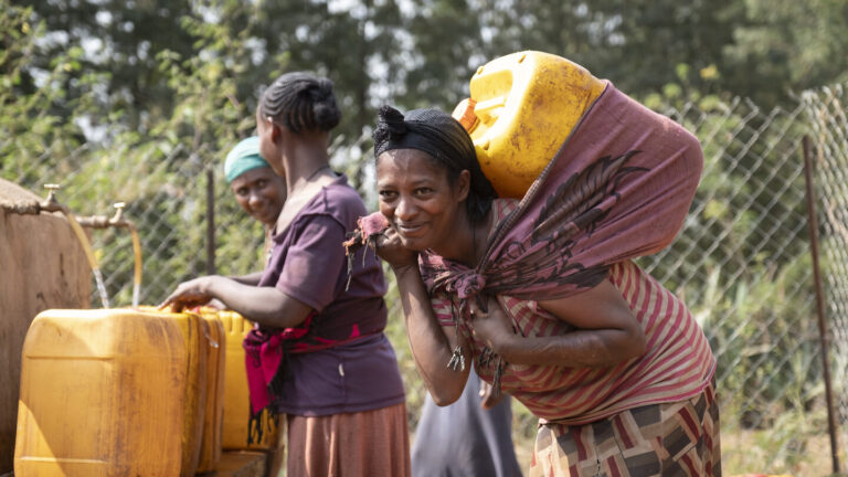Eine Frau im Vordergrund bindet sich einen vollen Wasserkanister auf den Rücken. Im Hintergrund sieht man zwei weitere Frauen mir ihren Kanistern.