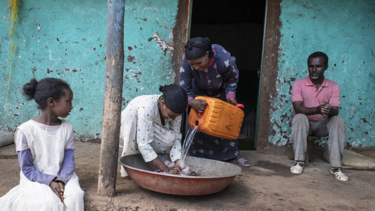 Zwei Frauen und ein Mädchen die zusammen Wäsche in einer Wasserschale waschen.