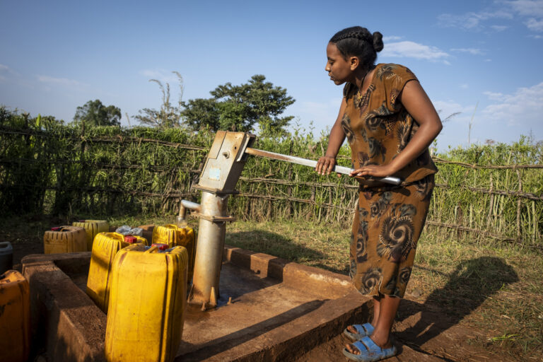 Eine junge Frau betätigt den Hebel an einem Brunnen und befüllt so ihre Wasserkanister.