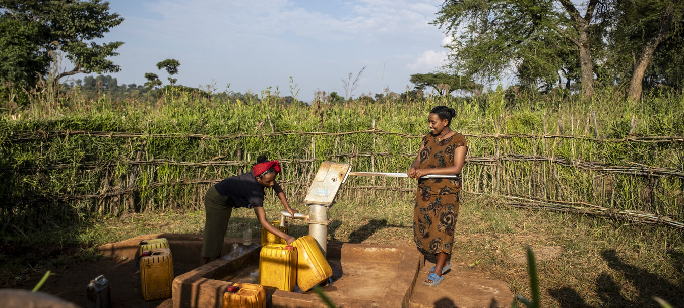 Zwei Personen stehen an einer Handpumpe für Wasser im Grünen.