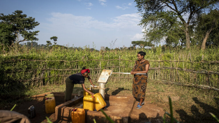 Zwei Personen stehen an einer Handpumpe für Wasser im Grünen.