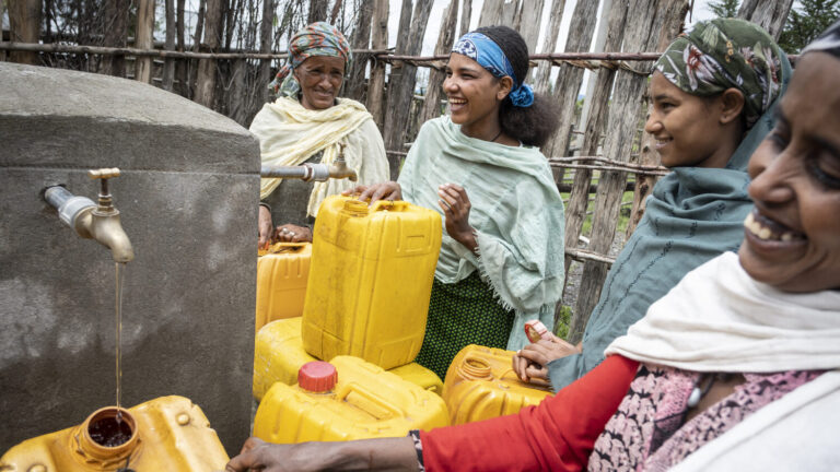Frauen stehen lachend um den Wasserbrunnen und befüllen Kanister.