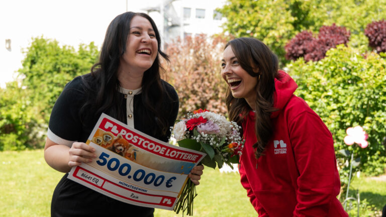 Zwei Frauen die lachen. Ein Mitarbeiter der Postcode Lotterie, die Gewinnerin, die ein Schild mit der Gewinnsumme hält.