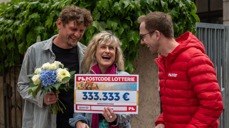 Drei Personen die lachen. Ein Mitarbeiter der Postcode Lotterie, zwei Gewinner, die ein Schiuld der Gewinnsumme halten.