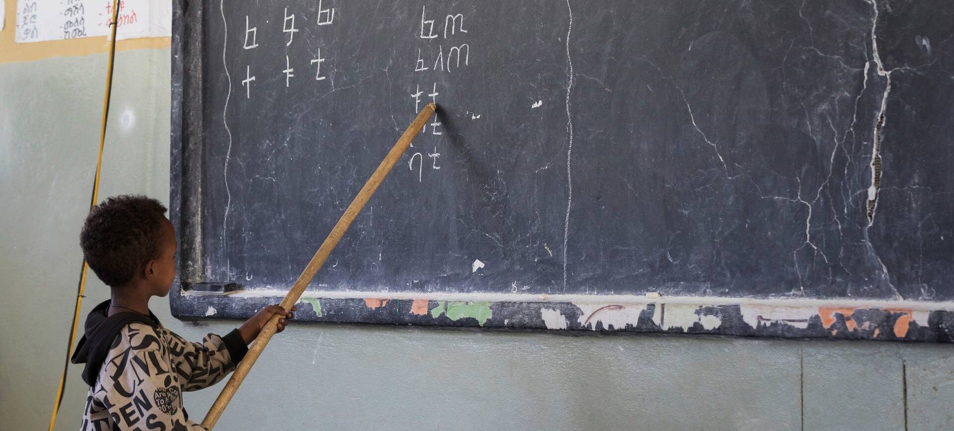 Junge zeigt mit Holzstab auf Buchstaben an einer Tafel