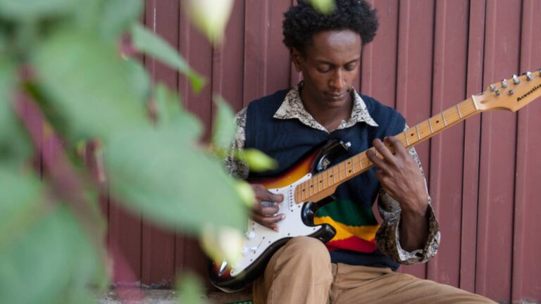 Mann mit E-Gitarre sitzt gegen eine Wellblechwand gelehnt und spielt