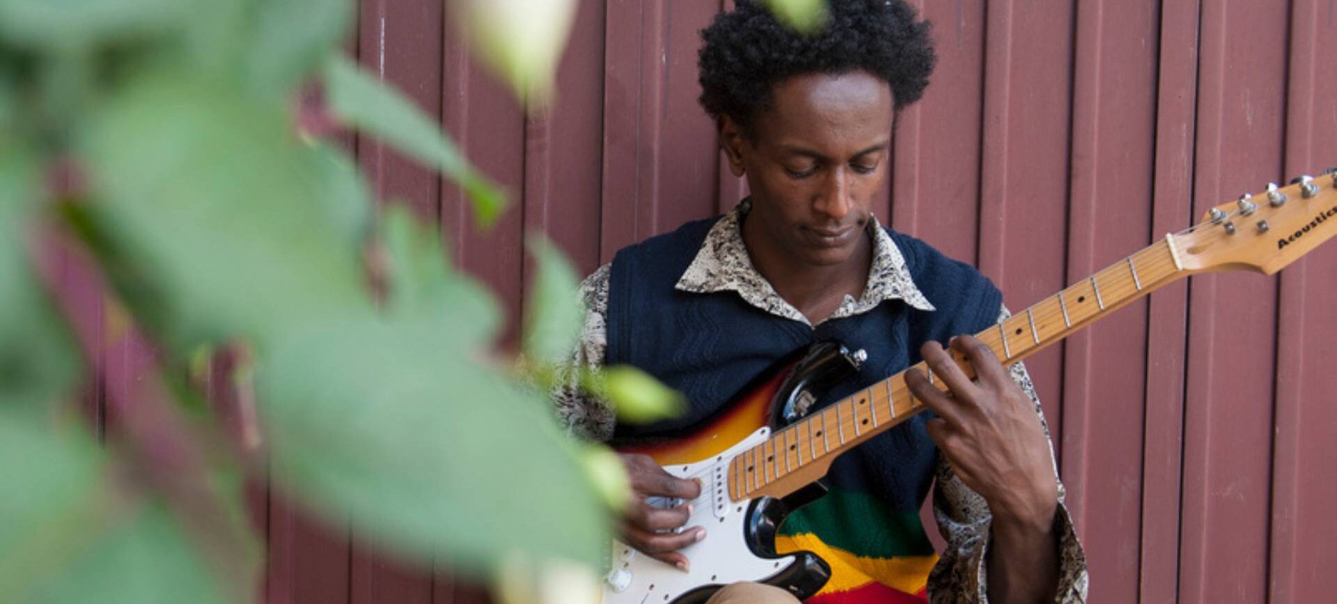 Mann mit E-Gitarre sitzt gegen eine Wellblechwand gelehnt und spielt
