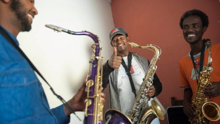 Drei Männer stehen nebeneinander mit Saxophonen in der Hand und lachen sich an
