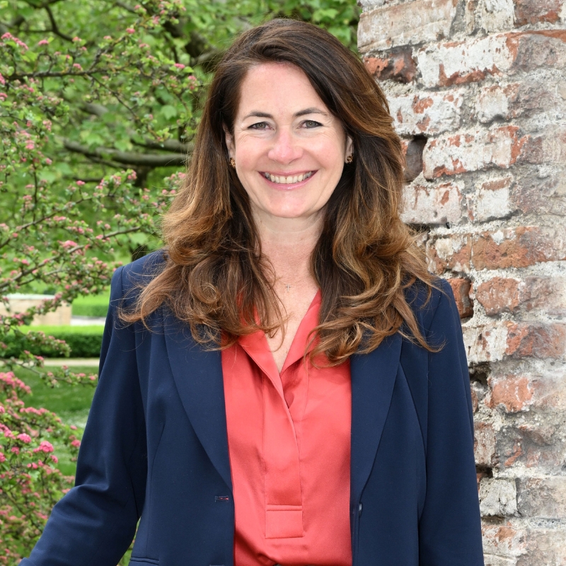 Eine professionell gekleidete Frau lächelt in die Kamera und steht vor einer Backsteinwand