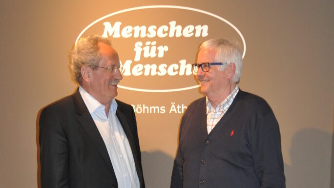 Dietmar Krieger und Christian Ude stehen vor einem Menschen für Menschen Logo und reden miteinander.