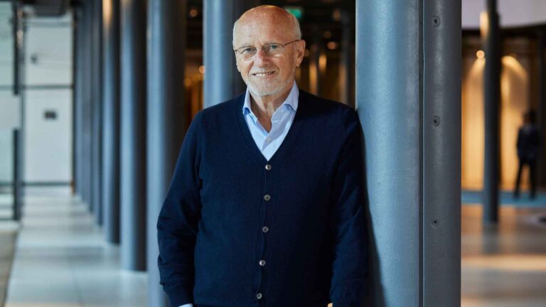 Dirk Roßmann, der Gründer der Drogeriemarktkette Rossmann, lehnt in einem Bürogebäude an eine Säule gelehnt und lächelt in die Kamera.