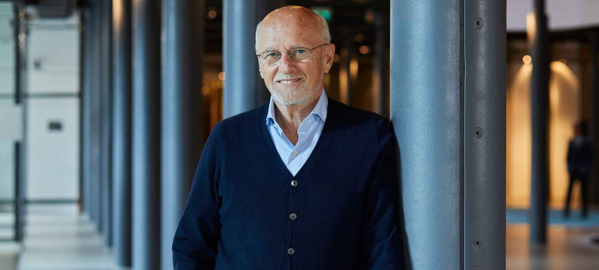 Dirk Roßmann, der Gründer der Drogeriemarktkette Rossmann, lehnt in einem Bürogebäude an eine Säule gelehnt und lächelt in die Kamera.